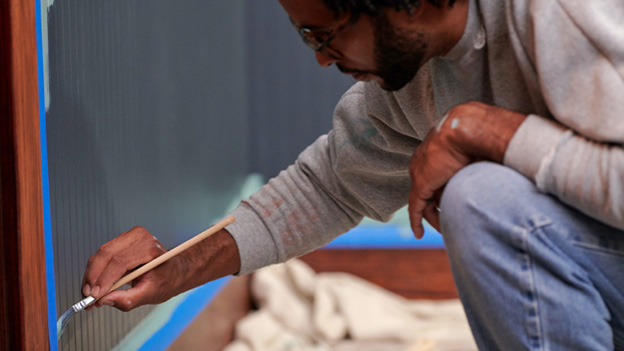 Person applying light-green color to wall with blue tape around a doorway with a drop cloth and a paint tray in background.