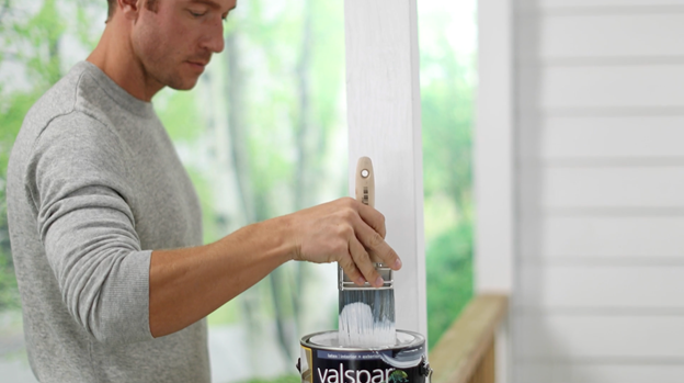 Person on porch dipping brush into can with an unfinished handrail in background.